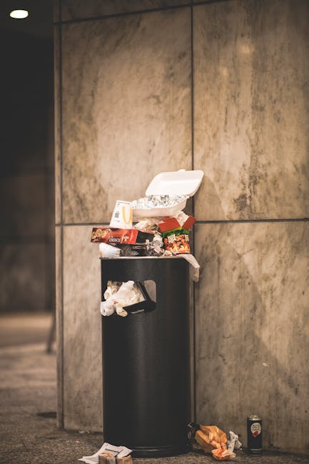 A black cylindrical rubbish bin with a partially open white plastic lid, positioned against a large beige tiled wall. The top of the bin is overflowing with various disposable food containers, paper w
